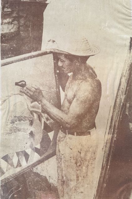 一名光着上身、戴草帽的男子用细笔在画布上作画，画布支在一副粗糙的木画架上，人站在画布与木板墙之间的狭窄空隙里。画布上，一条长着三角形牙齿的鱼已经占满了下半幅。棕褐色调的印刷复制品，网点清晰可见。