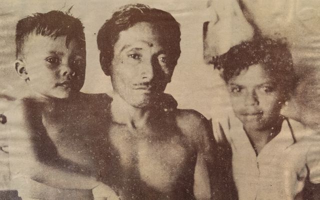 一名蓄细髭的年轻男子赤着上身，在两个孩子之间望向镜头：左边是一个头发竖起的小男孩，右边是一个穿浅色上衣的女孩，手搭在他肩上。棕褐色调的印刷复制品，网点清晰可见。