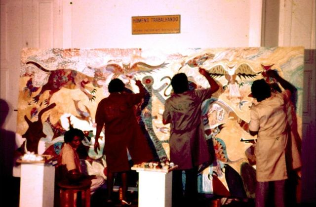 四名穿罩衫的画家背对镜头,同时在一幅宽大的奇幻生灵画板上作画;画板上方挂着写有 Homens Trabalhando 的牌子。左侧有人坐在矮凳上。
